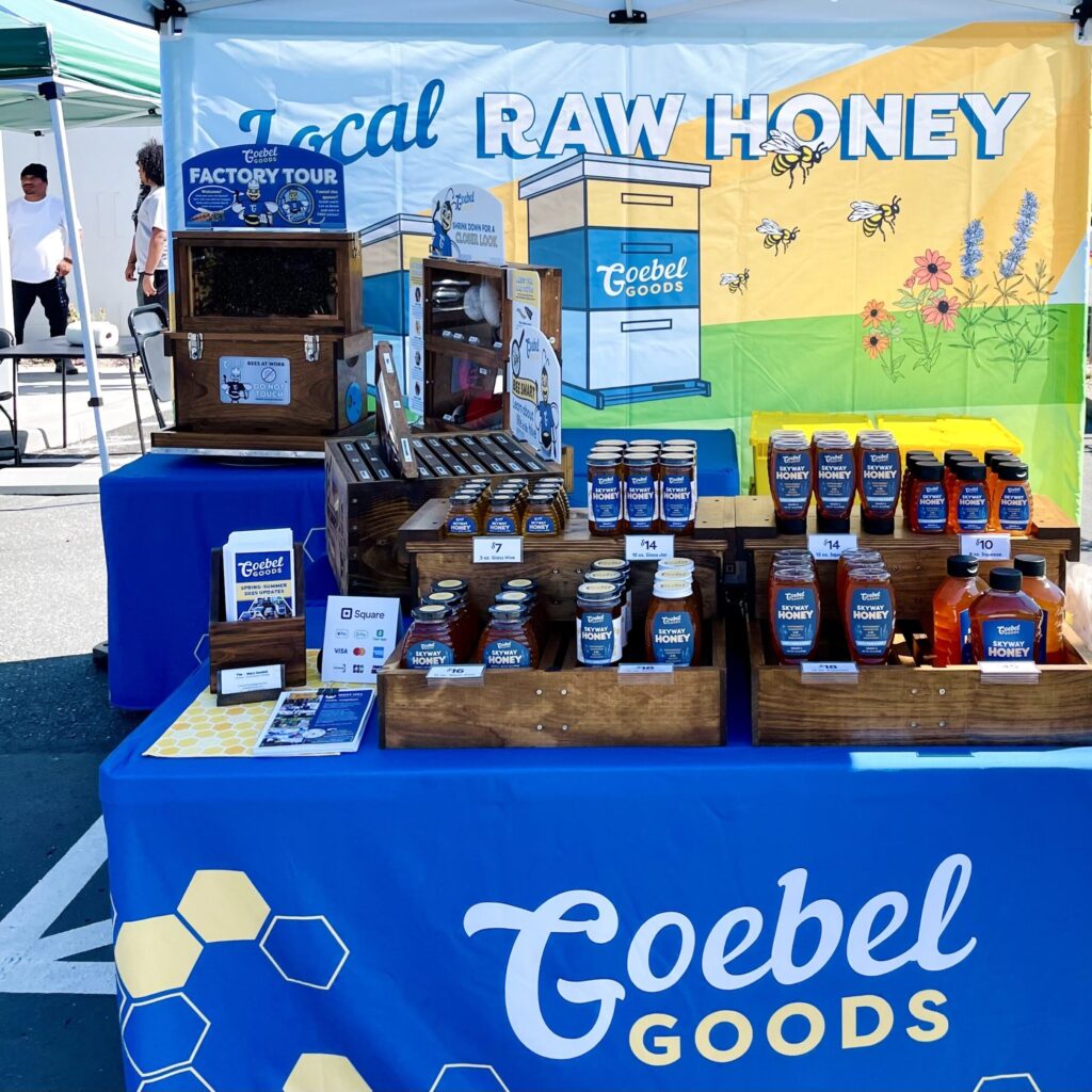 Table with blue tablecloth reads "Goebel Goods". There are neat rows of honey on the table and a banner in back that reads "Local Raw Honey".