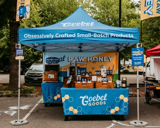 Goebel Goods booth setup at a farmers market. Blue canopy, branded tablecloths, honey display on table.